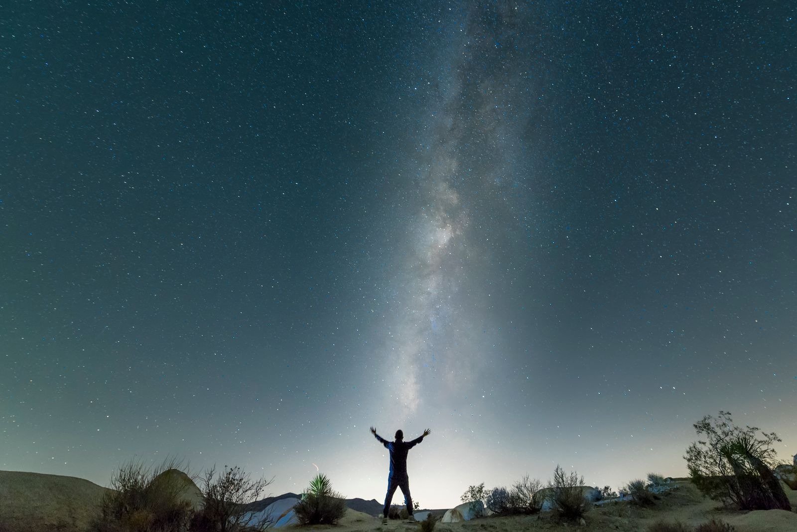 A silhouetted person with arms raised under the Milky Way in a desert night sky.