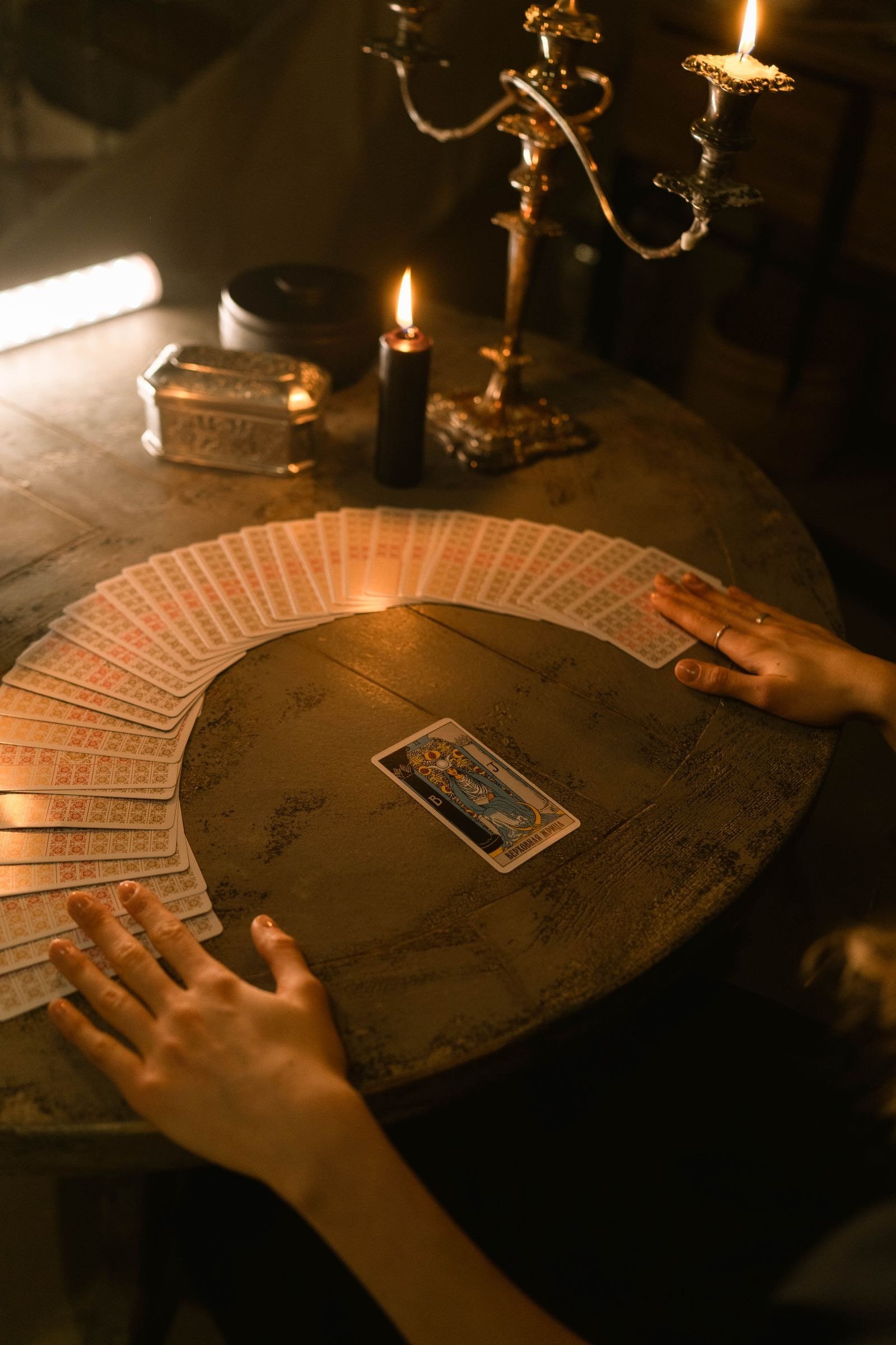 Hands fanning tarot cards in an arc on a round wooden table with candles and a candelabra.
