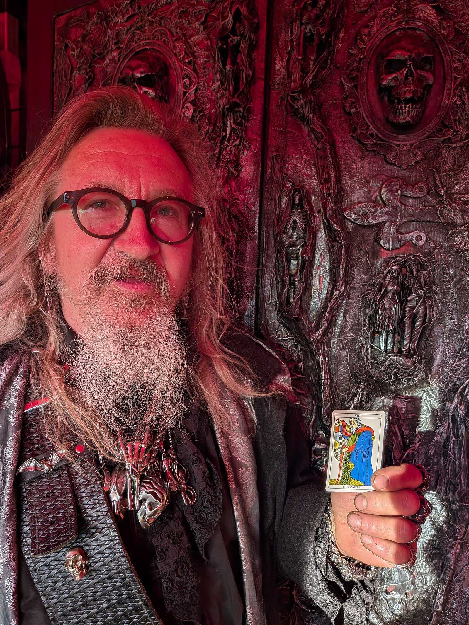A bearded man holding up The Hermit tarot card in front of an ornately carved dark-red door decorated with skulls.