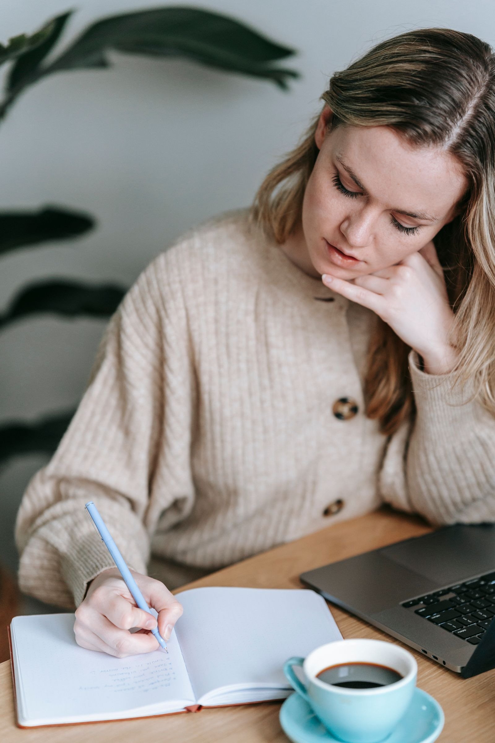A woman writing in a notebook at a table with a laptop and a cup of coffee.
