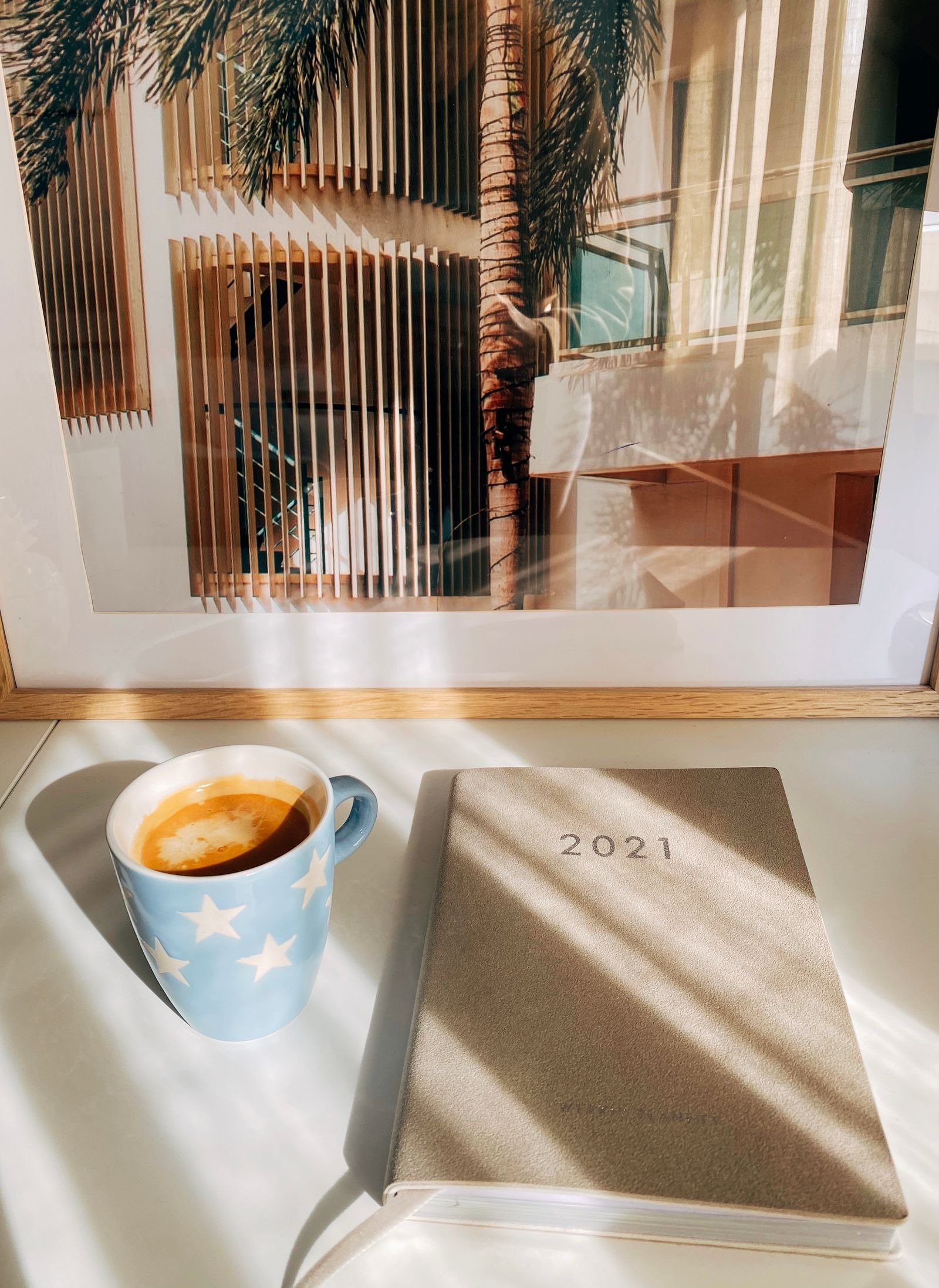 A closed planner beside a star-patterned coffee mug on a sunlit desk.