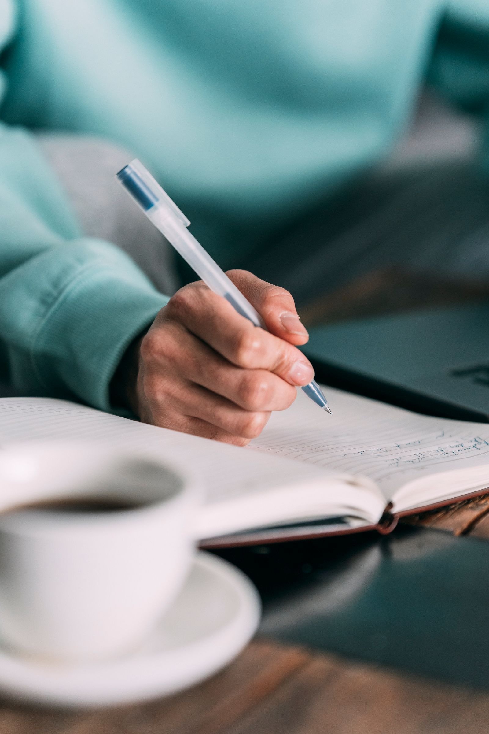 A close-up of a hand writing in a notebook with a pen, a coffee cup nearby.