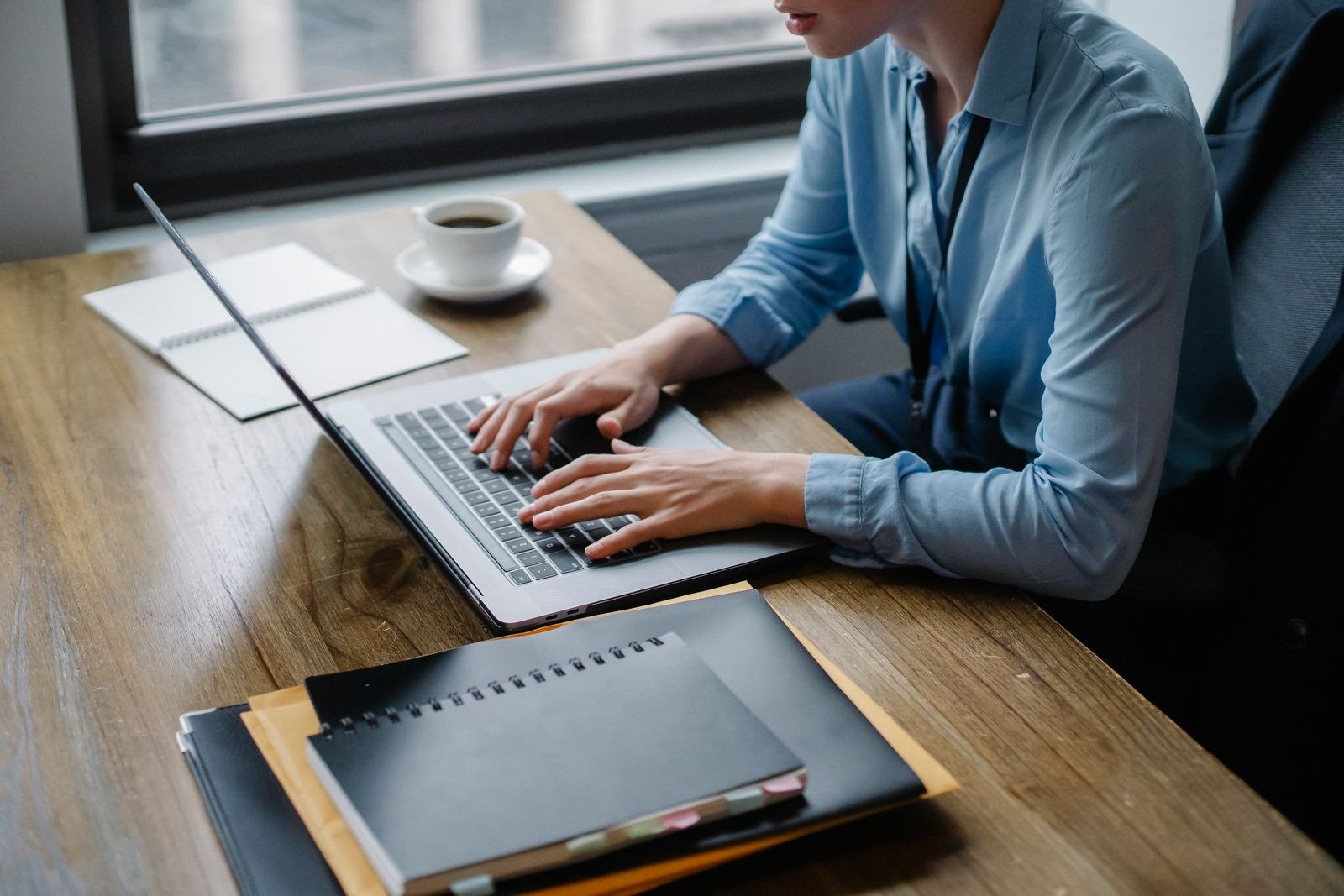 A person in a blue shirt typing on a laptop at a wooden desk, with a coffee cup and notebooks nearby.