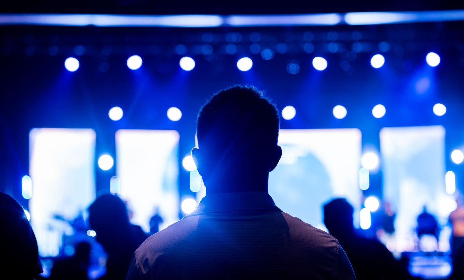 A silhouetted audience member facing a bright blue-lit stage with spotlights.