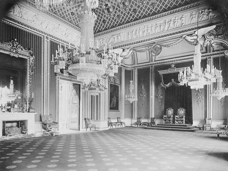 A 1914 black-and-white photograph of the throne room at Buckingham Palace, with chandeliers and ornate thrones on a raised dais