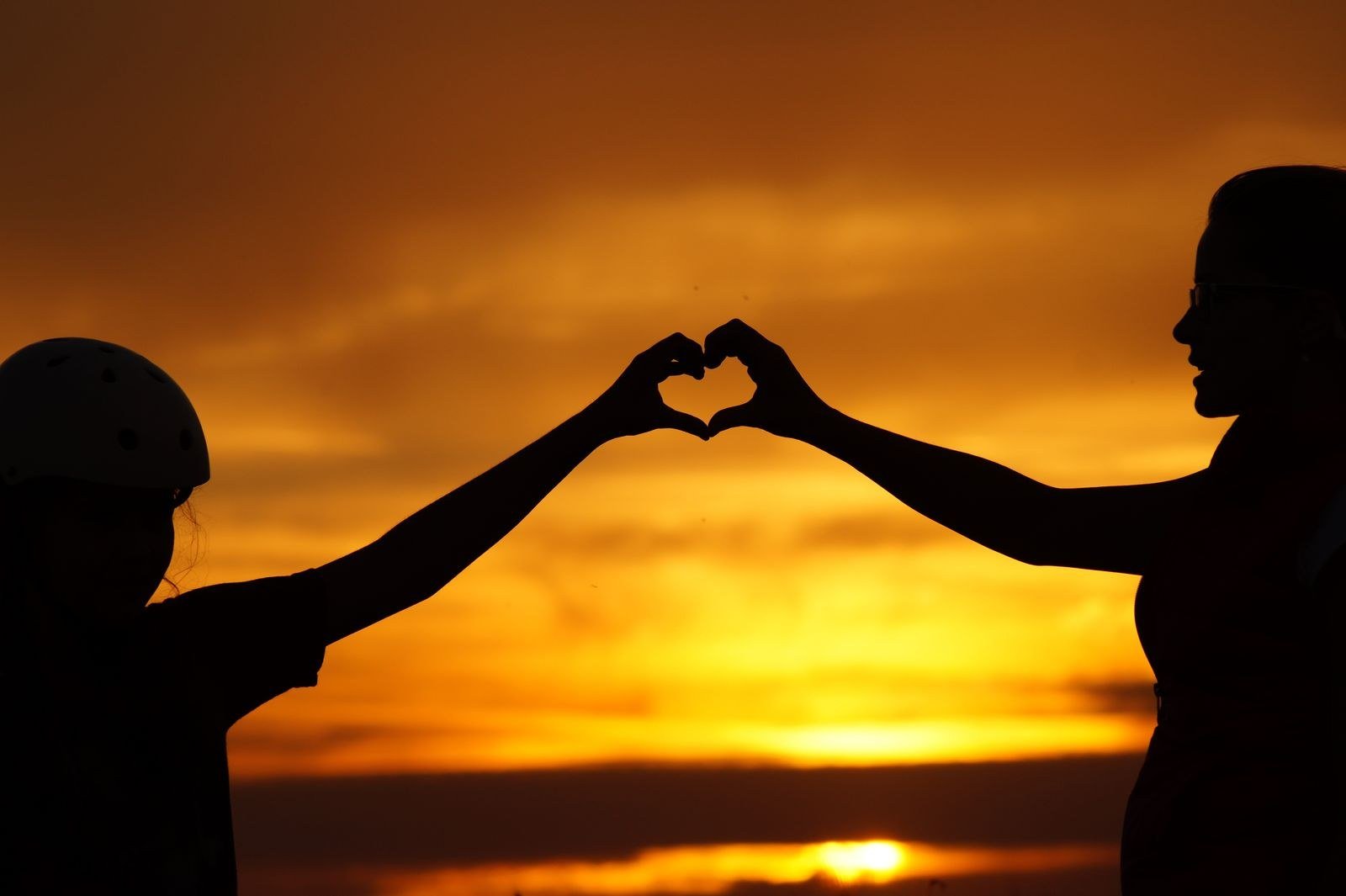 Two people in silhouette forming a heart shape with their hands against an orange sunset.