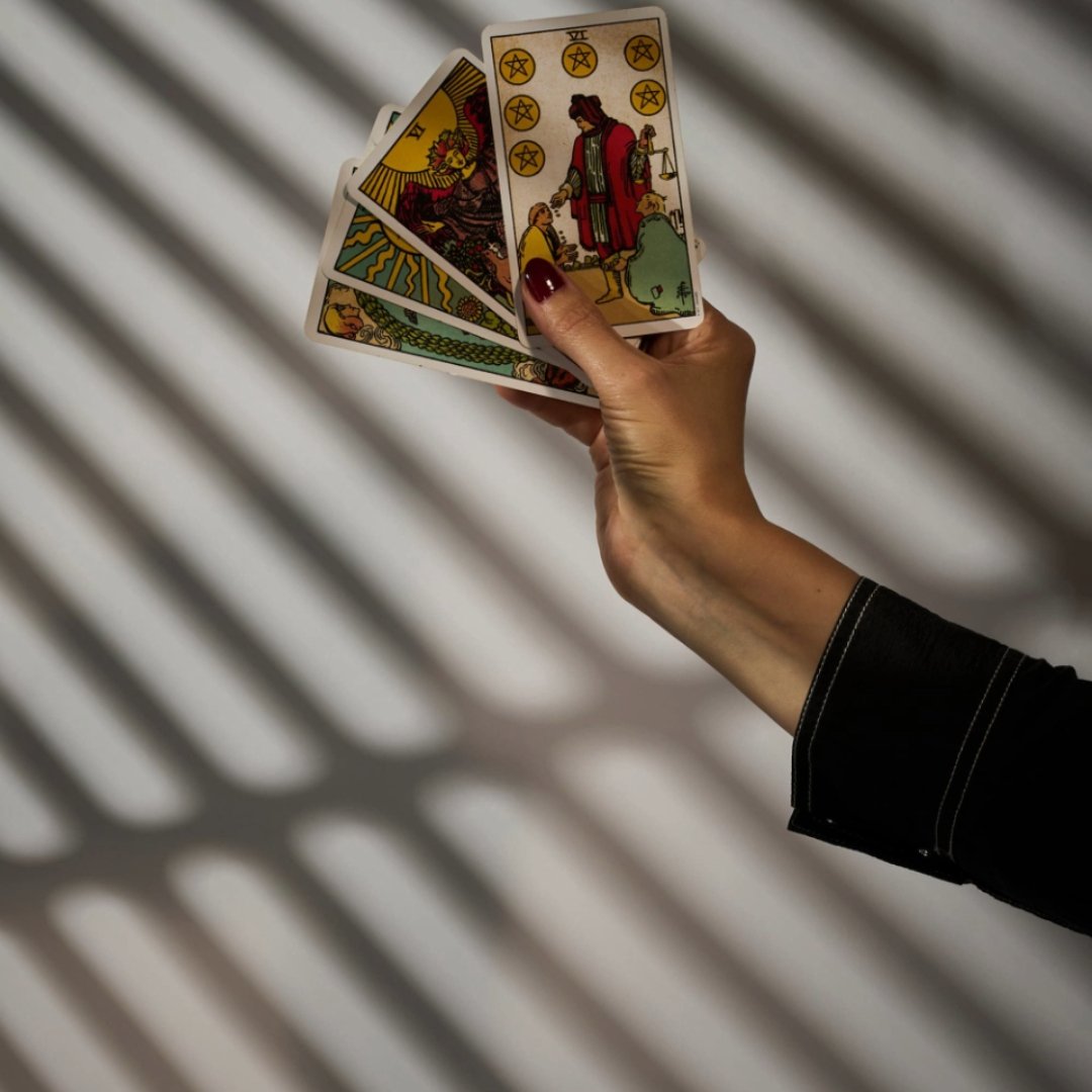 A hand holding several Rider-Waite-style tarot cards against striped window light.