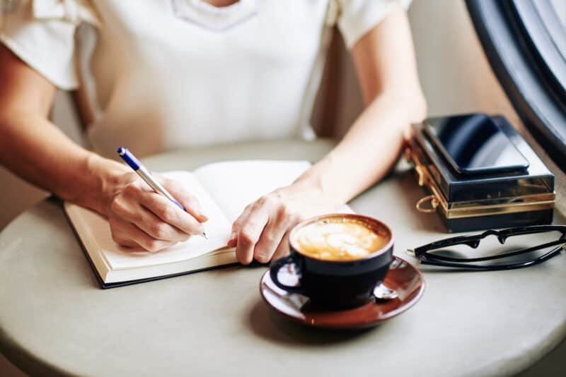 A woman writing in a journal at a table in the morning.