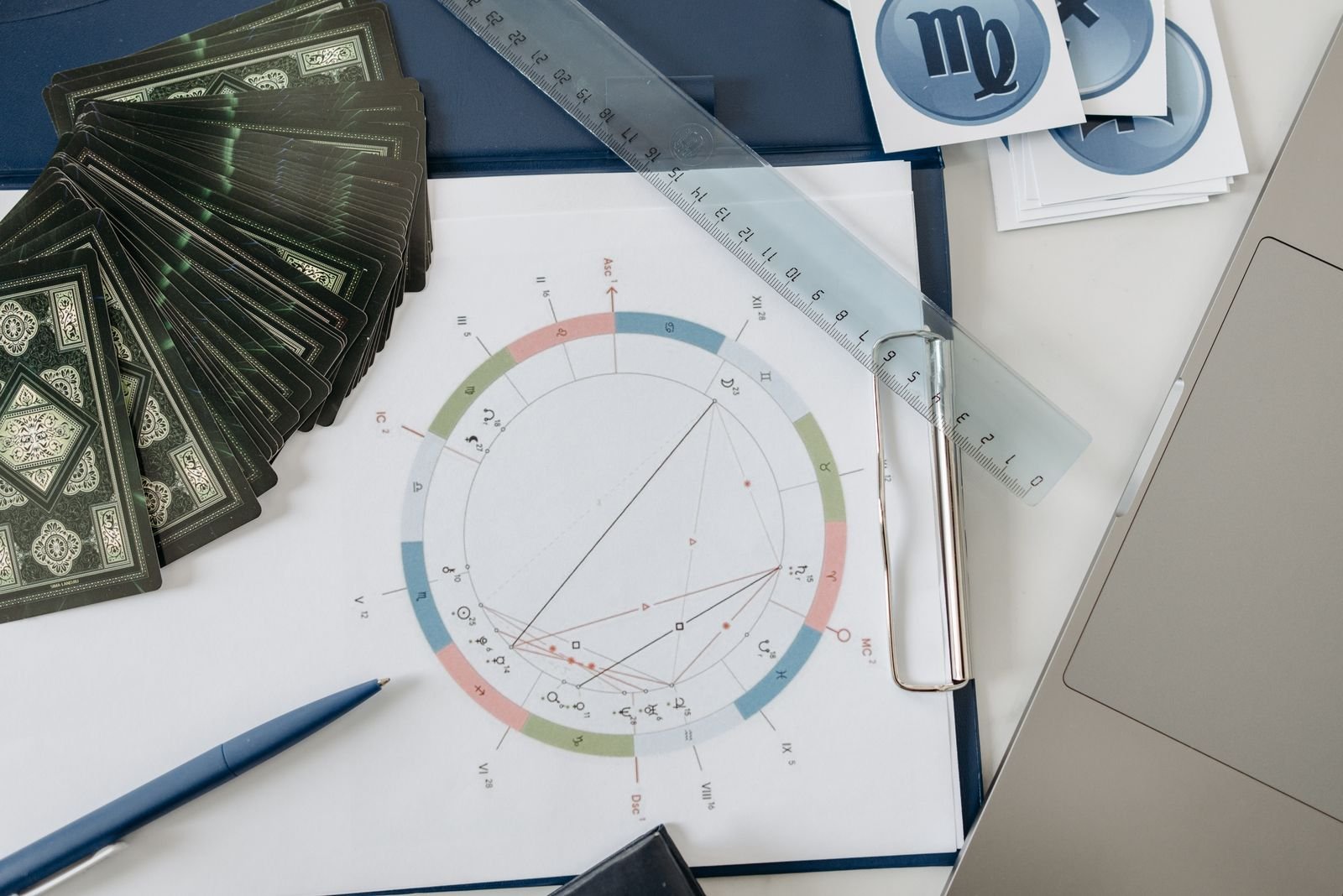 A flat lay of an astrology study setup: a printed natal chart on a clipboard with a ruler and pen, fanned tarot cards, and Virgo symbol cards beside a laptop.