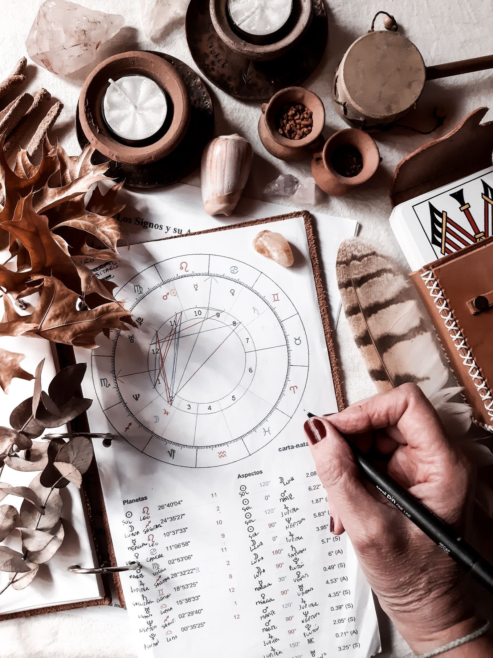 A hand holding a pen over a printed natal chart with handwritten planet and aspect tables, surrounded by candles, dried leaves, shells, crystals, a feather, and tarot cards in a leather case.