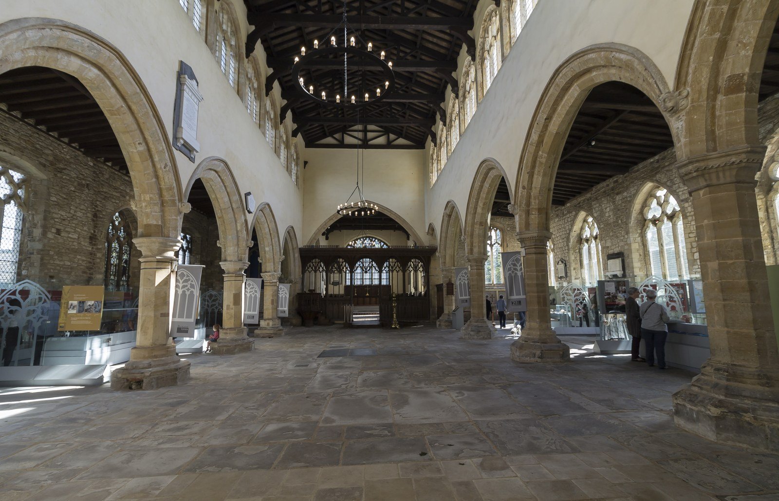 Sunlit interior of a stone church with arched columns, hanging chandeliers, and a few visitors