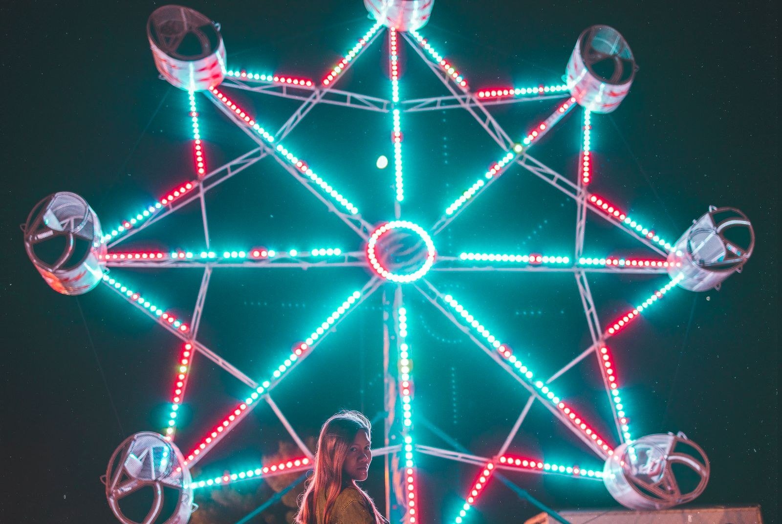 Ferris wheel lit with red and teal lights at night, with a woman standing in front.