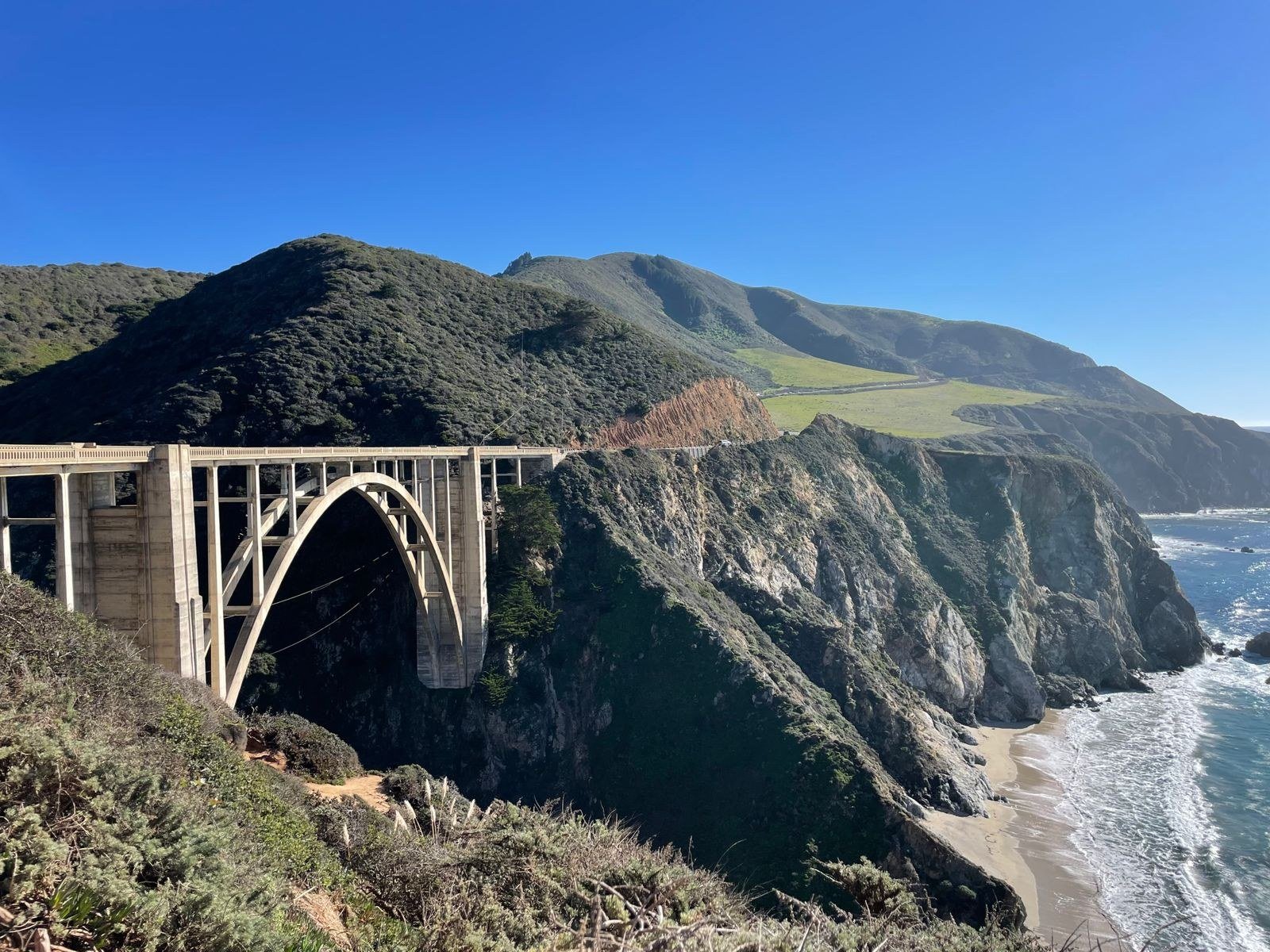 Concrete arch bridge spanning a coastal canyon with mountains and ocean below.