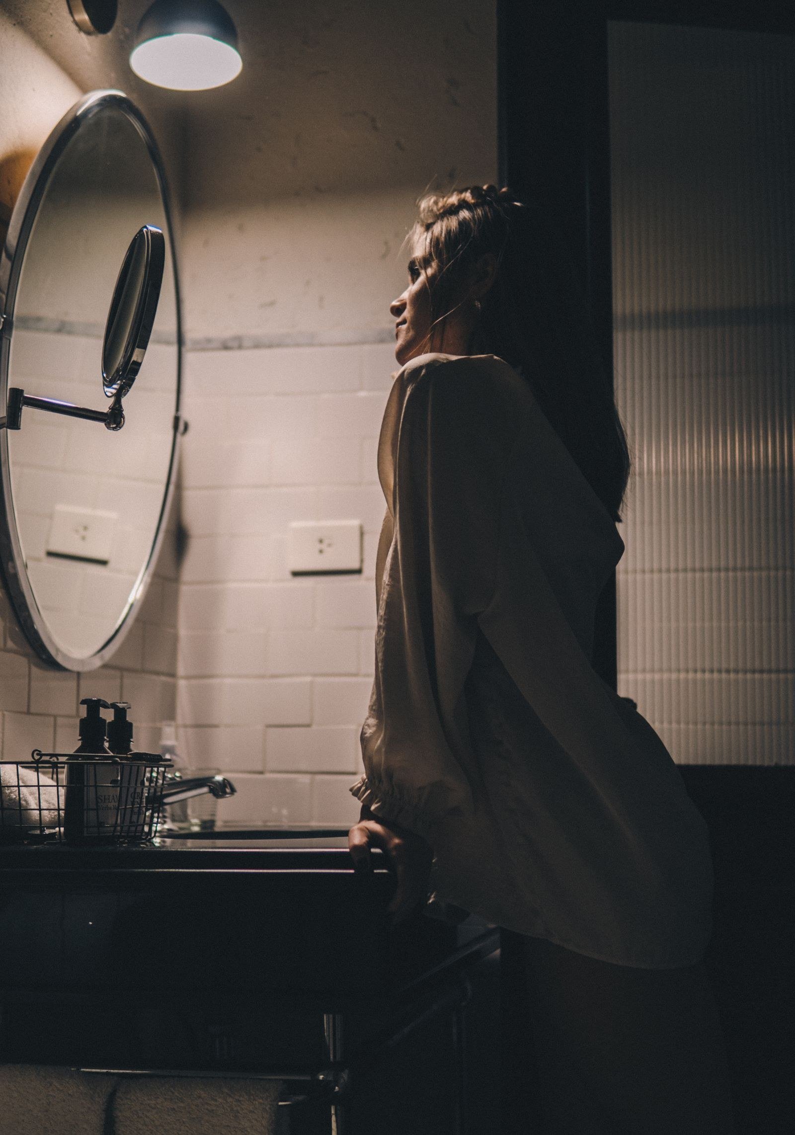 A woman studying her reflection in a round mirror in soft window light, one hand touching her hair.
