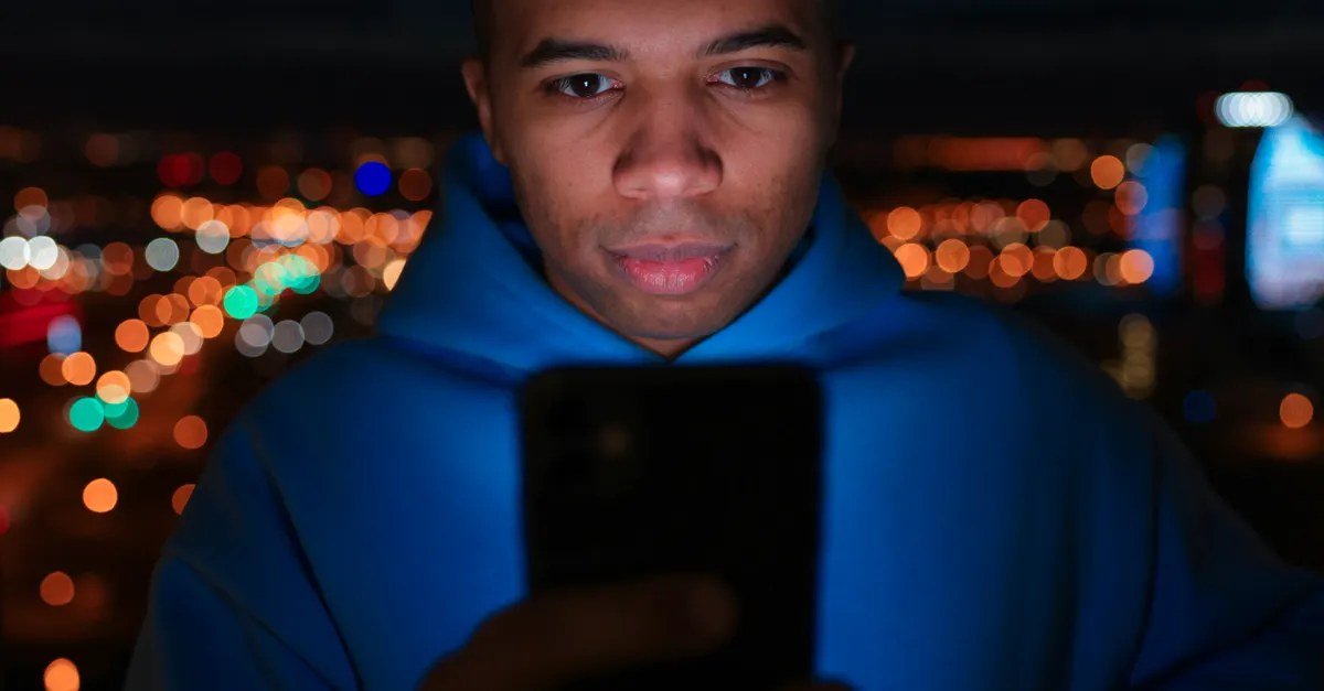 Person in a blue hoodie looking at a smartphone at night with blurred city lights behind