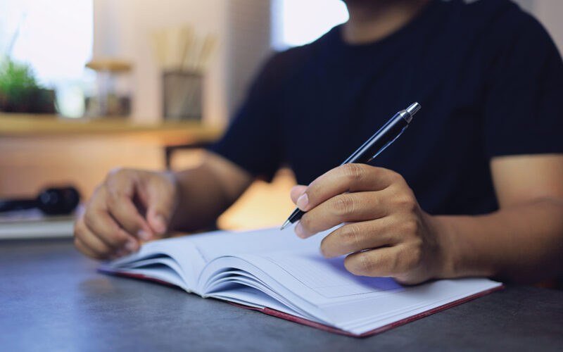 A person writing in a notebook with a pen at a desk